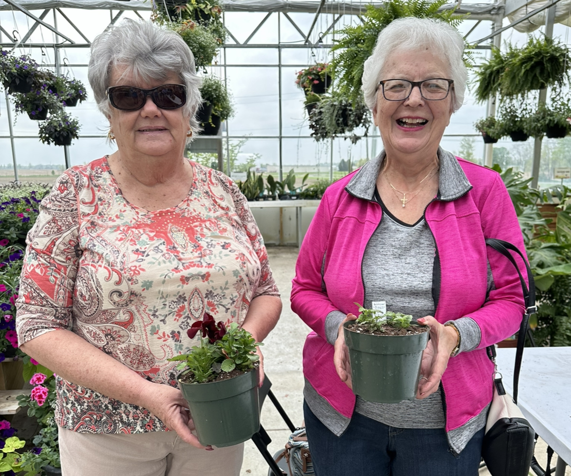 Two members standing inside a greenhouse holding flowers they planted in a pot. 